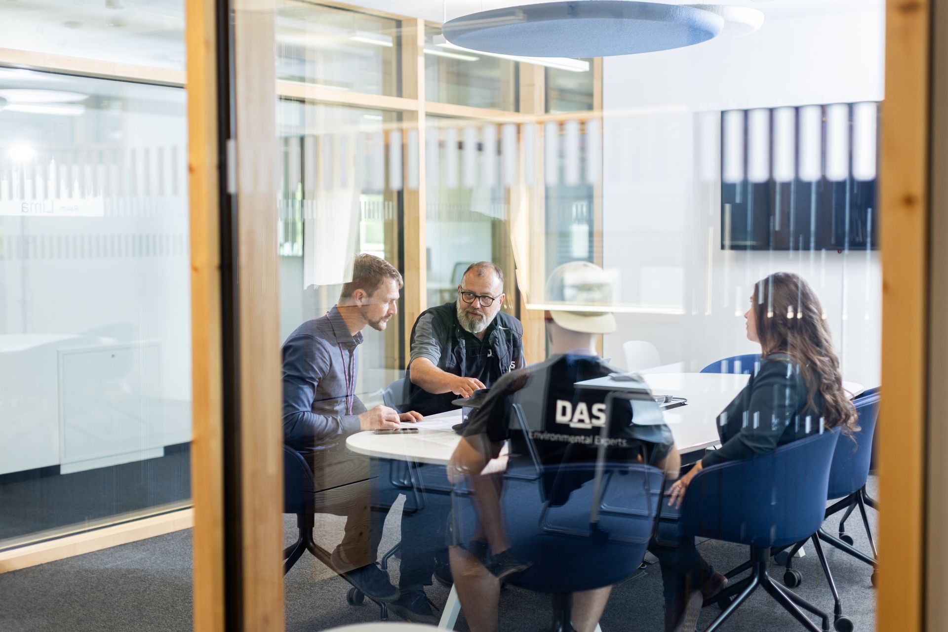 A group of four people sit around a table in a modern glass-walled office, engaged in a collaborative meeting, with office furniture and a digital screen visible in the background.