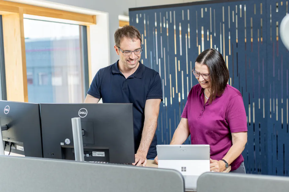 Service Team A man and a woman work smiling at a modern workstation with computers and a tablet, surrounded by stylish blue acoustic panels that create a productive office environment.