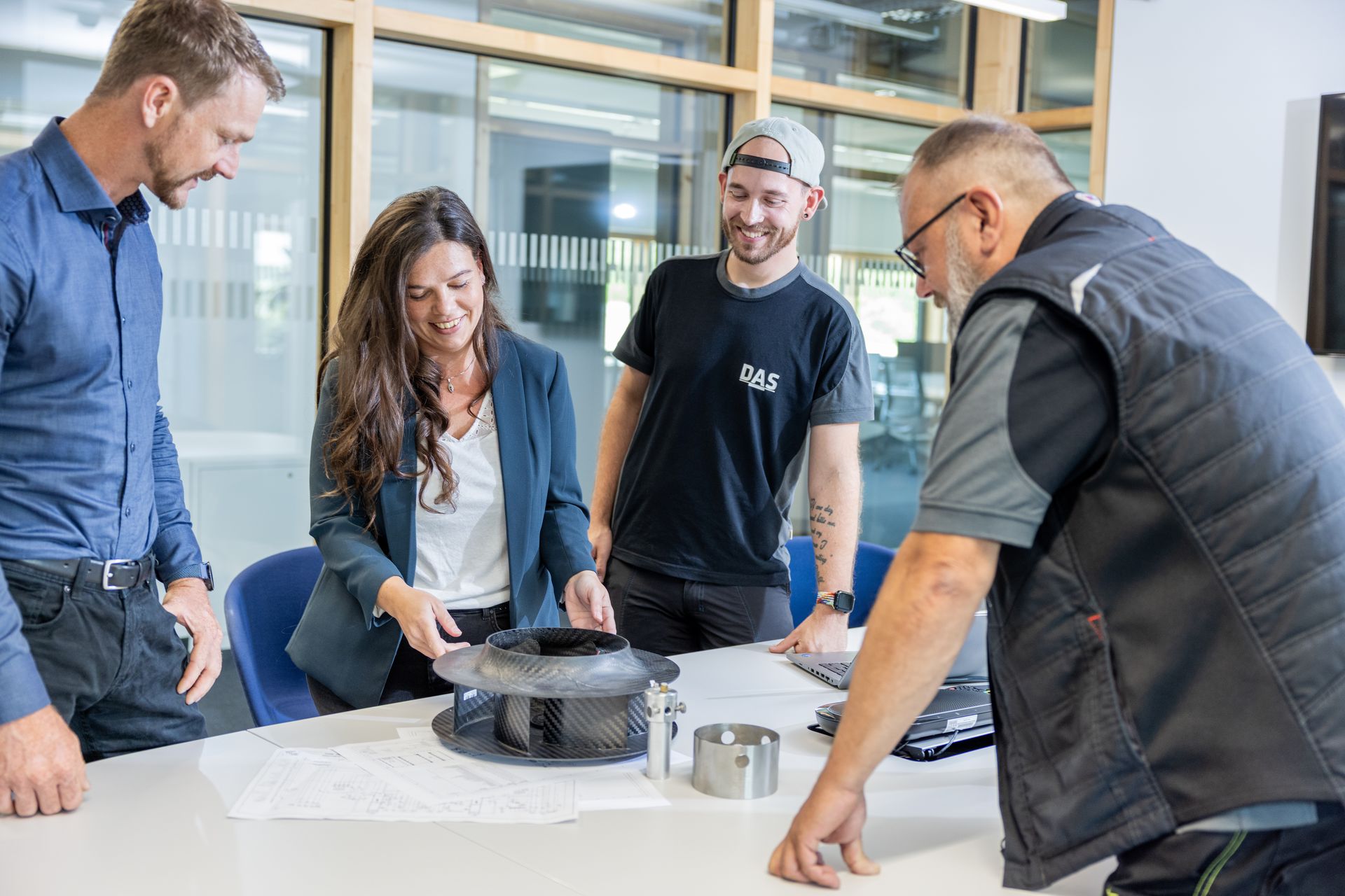 A group of four people stands around a table examining mechanical parts and technical drawings, engaged in a collaborative discussion in a modern office setting.