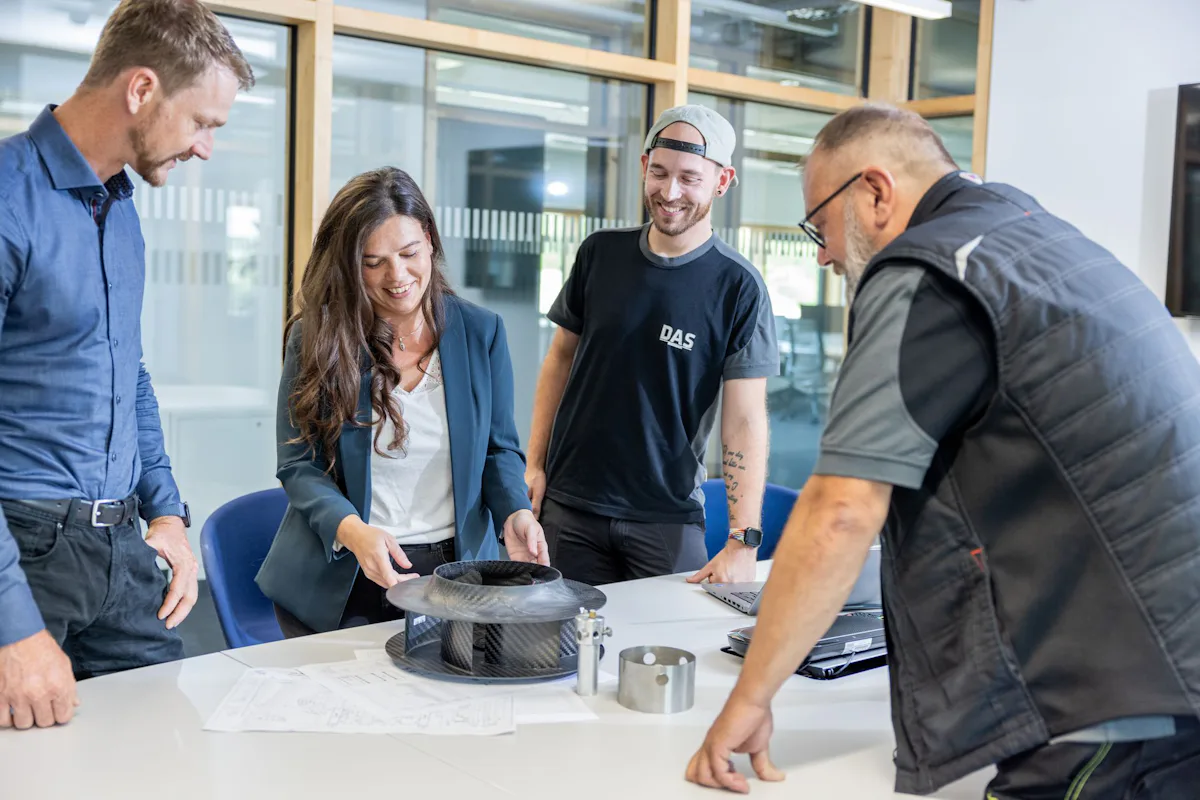 Mitarbeiter Meeting A group of four people stands around a table examining mechanical parts and technical drawings, engaged in a collaborative discussion in a modern office setting.