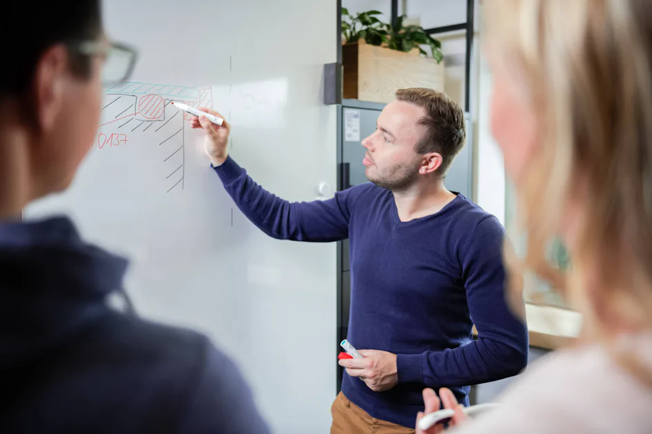 Planning and Concepts A man draws complex diagrams on a whiteboard with a red marker while two people listen attentively.