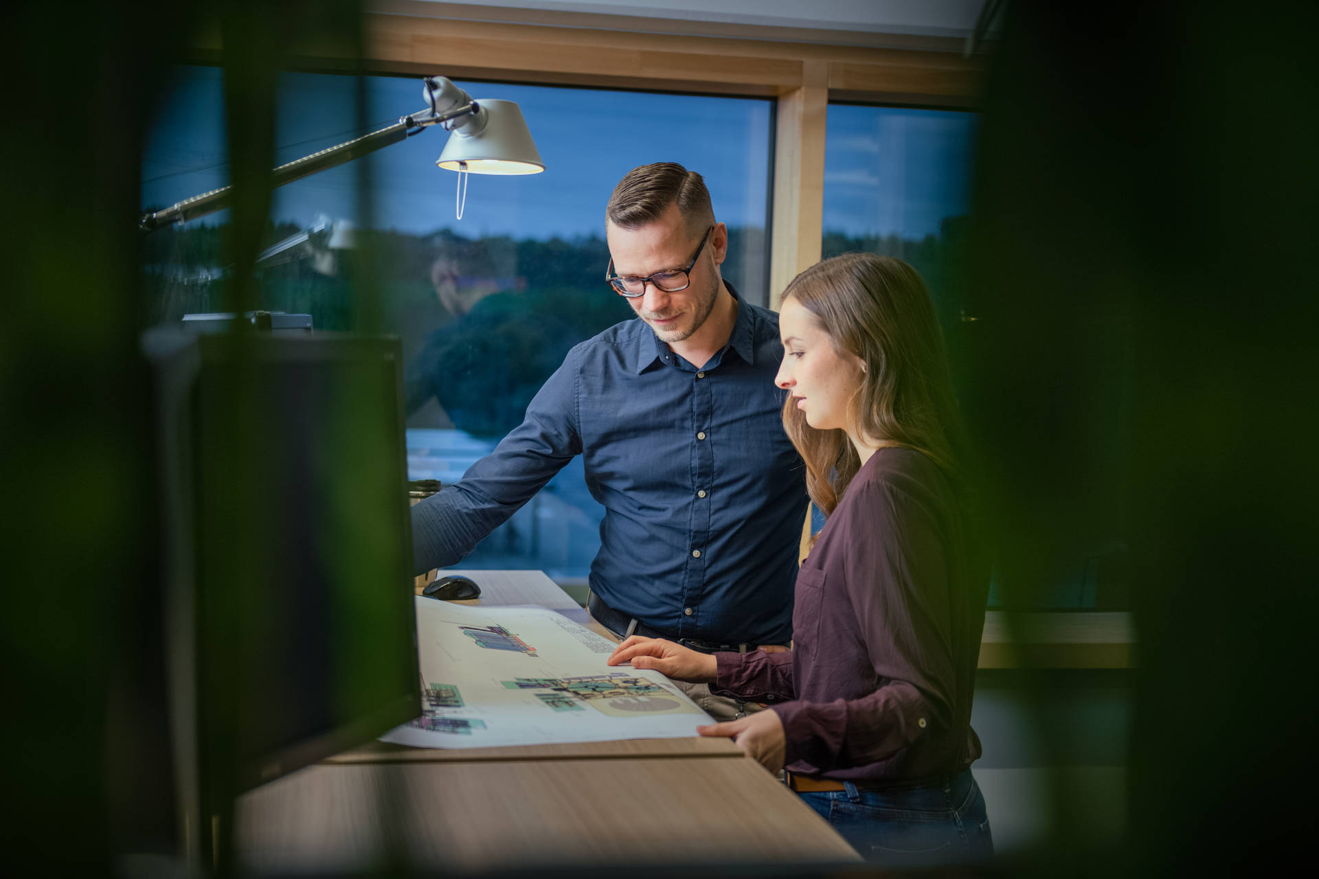 A man and a woman are looking at plans together on a table in a modern office with subdued lighting.