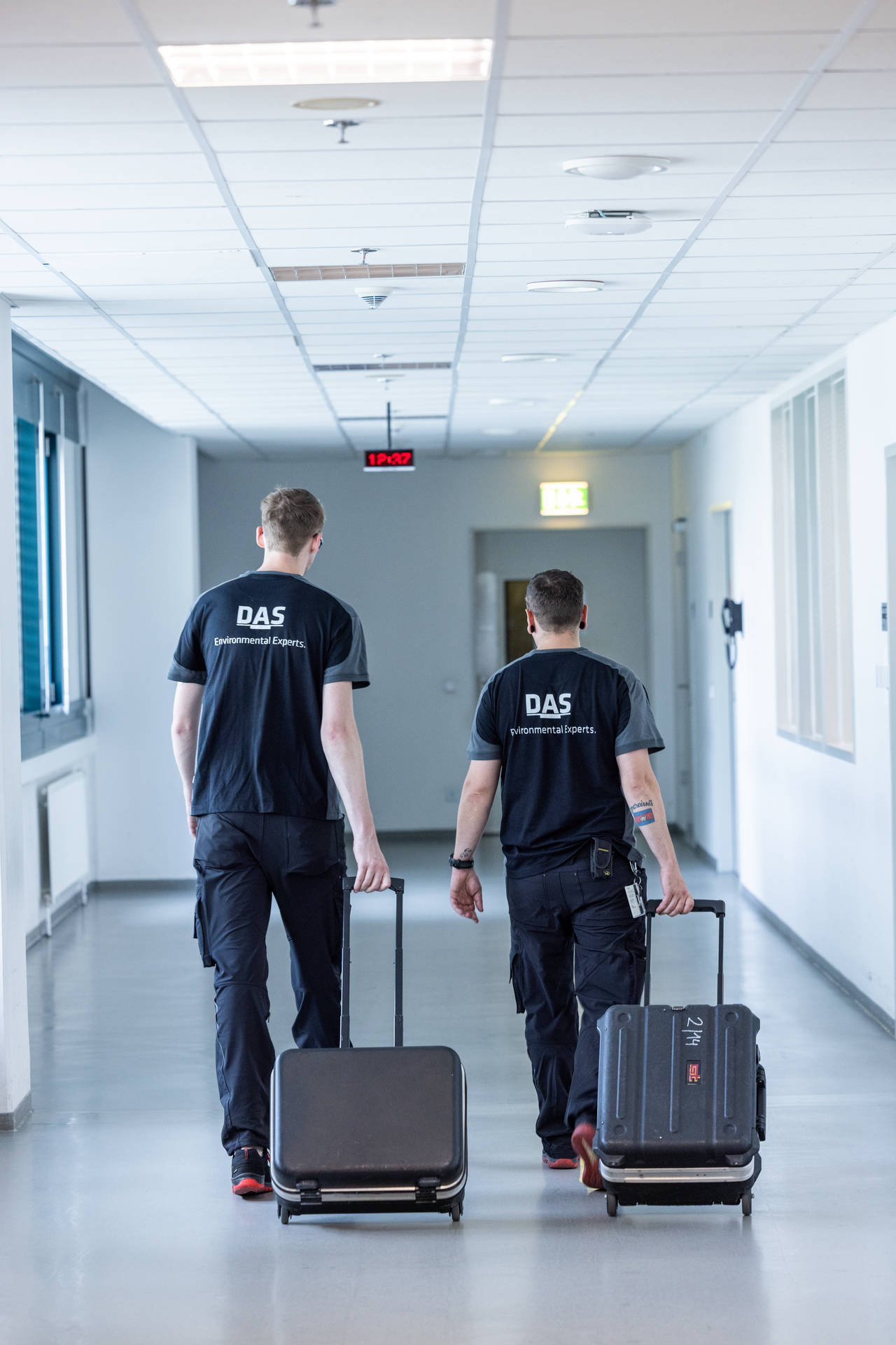 Two service employees wearing DAS uniforms walk down a brightly lit hallway, each pulling a suitcase.