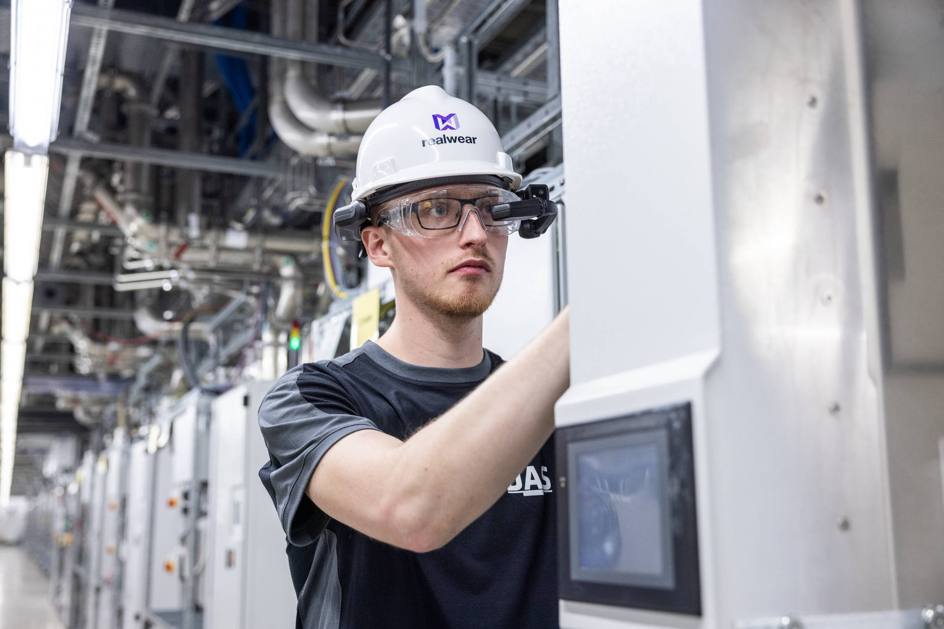 A worker wearing a hard hat and smart glasses operates machinery in an industrial facility filled with intricate piping and electrical equipment.