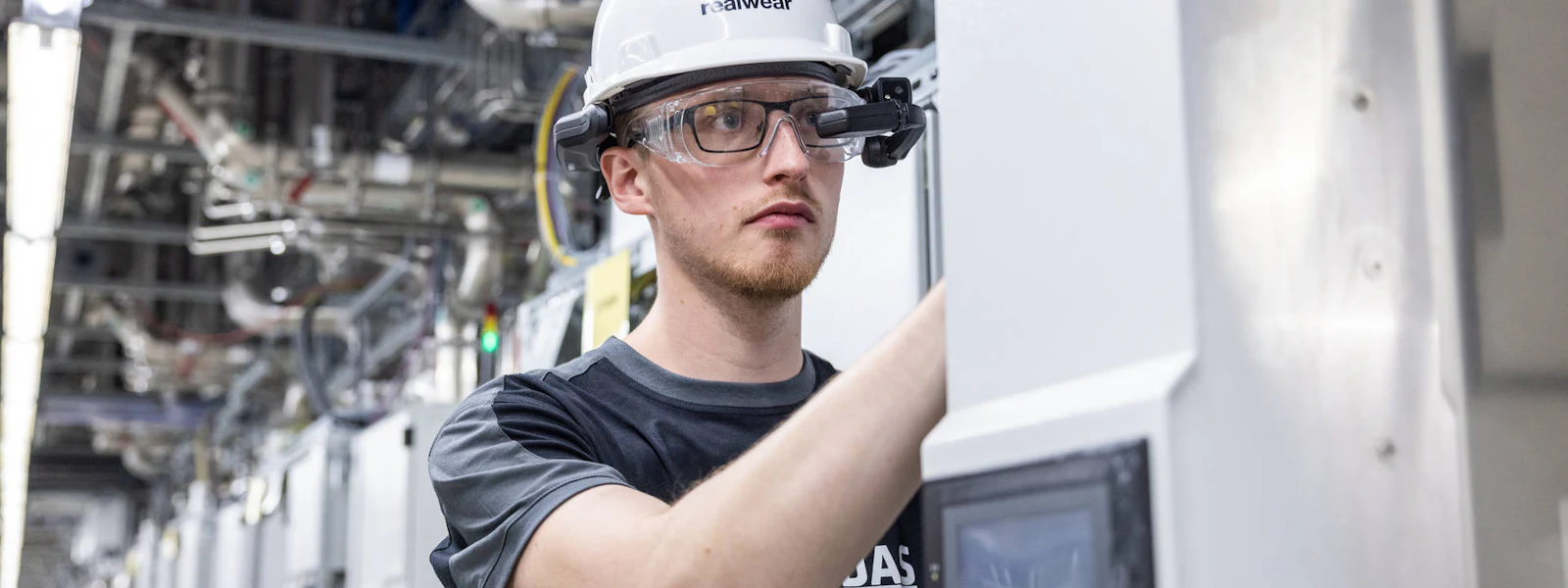 DAS employee working on abatement system 申し訳ありませんが、画像に表示されている人物について特定することはできません。ただし、画像の内容に基づいて代替テキストを作成しました。
ヘルメットとスマートグラスを着用した作業員が工場の制御パネルを操作している。