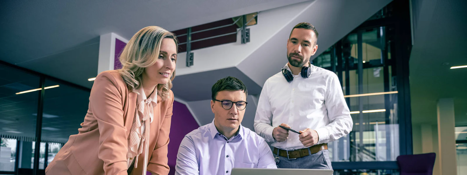 Drei Geschäftspersonen diskutieren konzentriert an einem runden Tisch mit einem Laptop und Kaffeetasse, in einem modernen Büro mit Glas und Treppenelementen im Hintergrund.