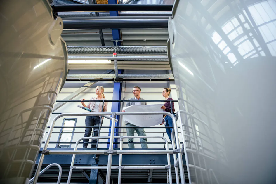 Besprechung Mitarbeitende Three people stand on a raised platform in an industrial facility and discuss a large construction plan, surrounded by metal structures; facility meeting.