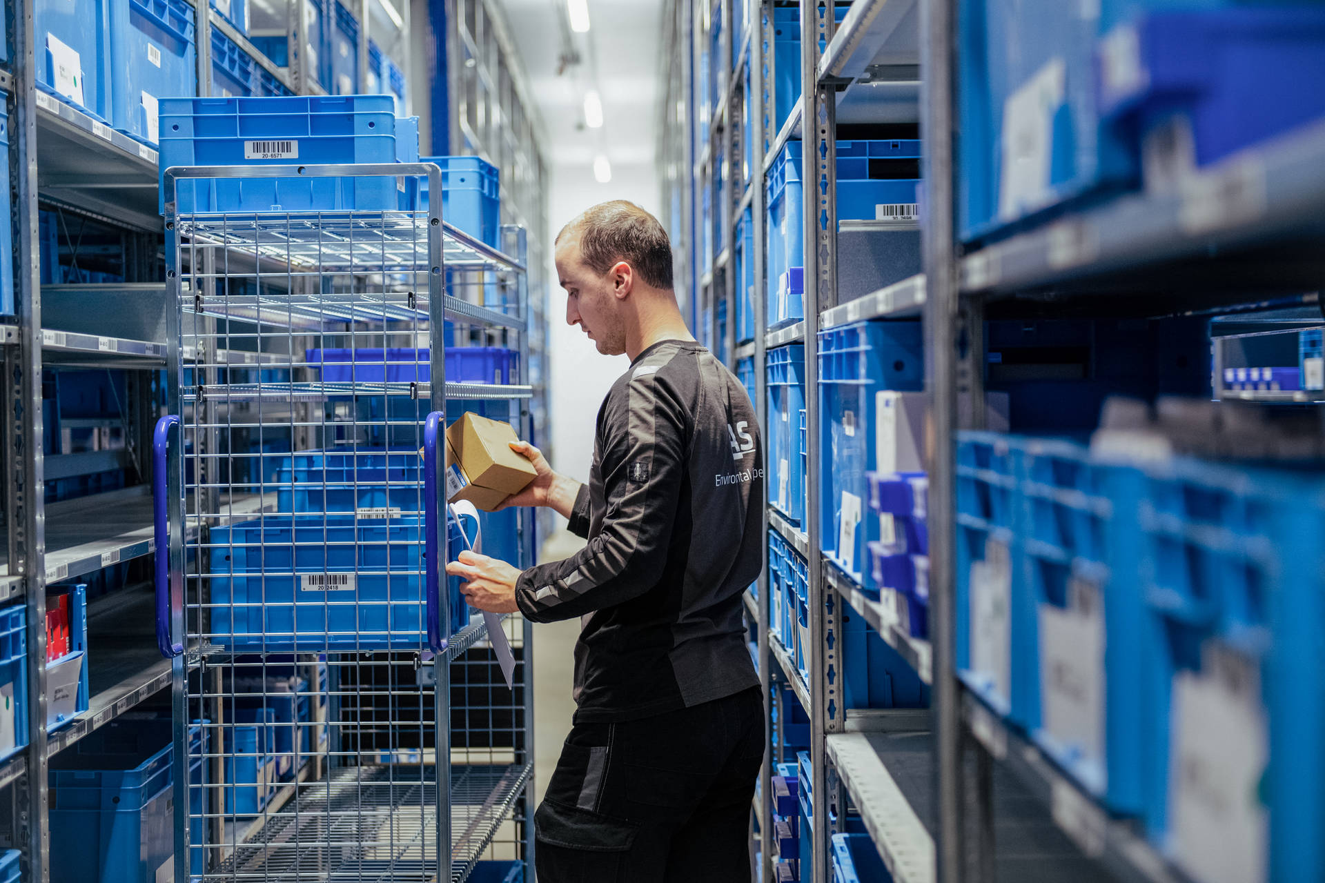 A worker in a warehouse sorts parcels on a blue shelf, surrounded by blue plastic crates, in a well-organised industrial environment.