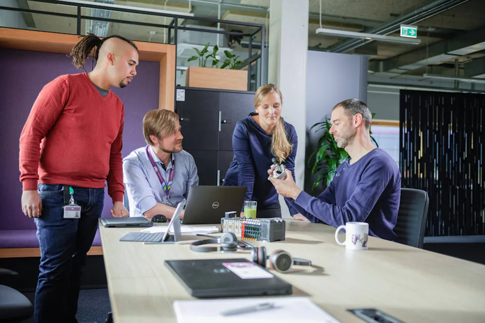 A group of four colleagues engage in a discussion around a conference table, examining a mechanical component, with a laptop and office equipment in the contemporary workspace. A group of four colleagues engage in a discussion around a conference table, examining a mechanical component, with a laptop and office equipment in the contemporary workspace.
