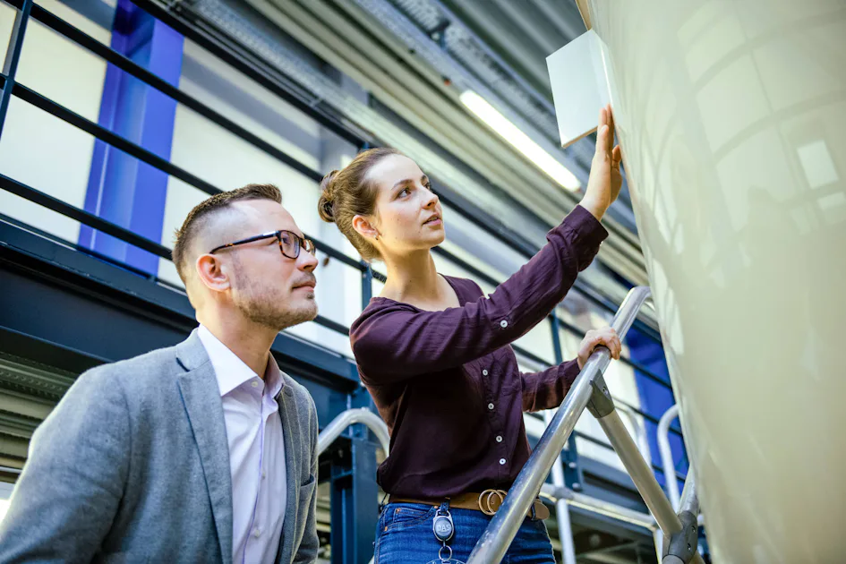 DAS Employees 587 Two individuals in business attire observe and take notes on a large industrial structure inside a modern facility, showcasing a collaborative work environment in a tech-oriented setting.