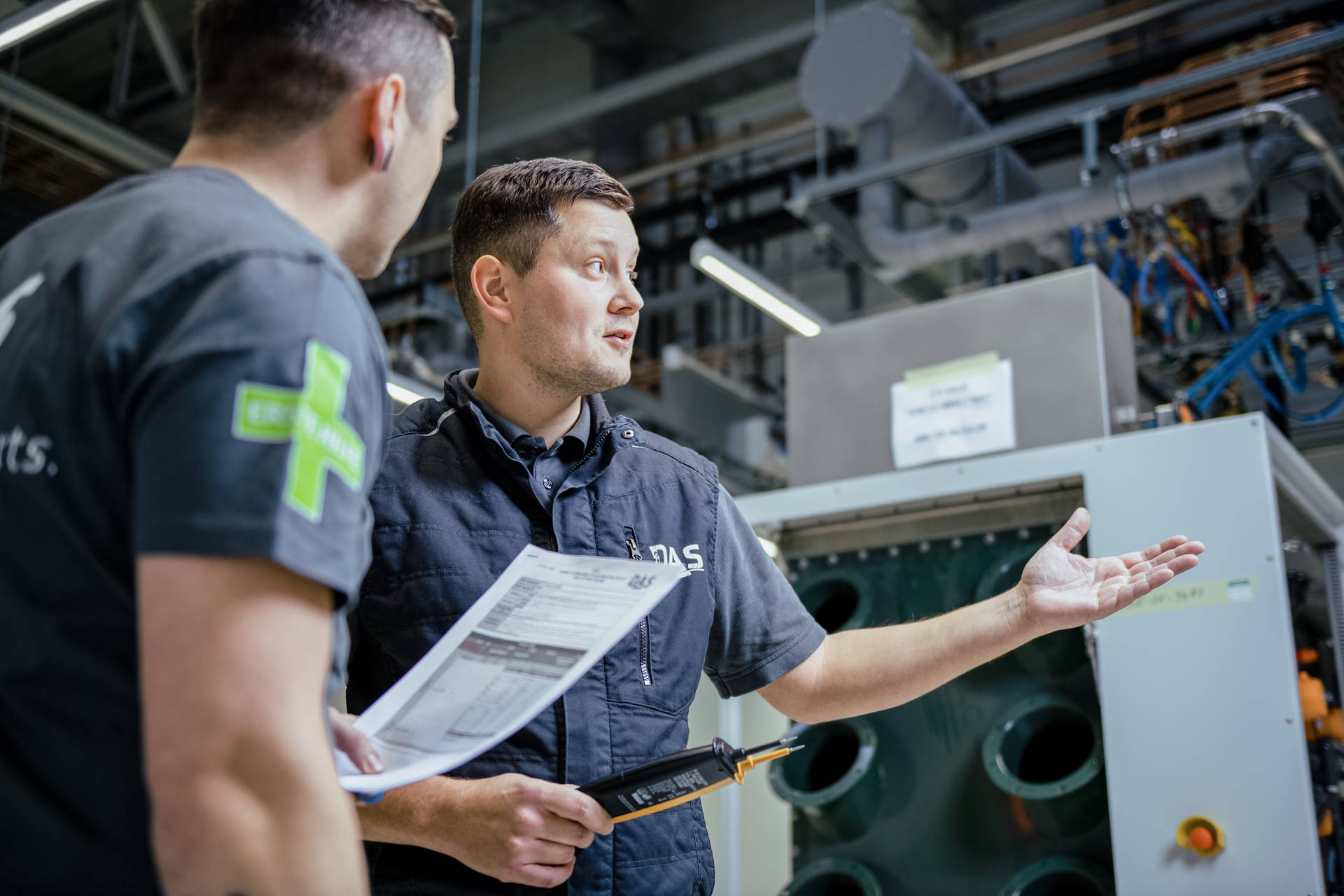 Two men in an industrial setting are conversing in front of a large machine unit, with one holding a document and the other making an explanatory hand gesture, surrounded by pipes and structural cabling.