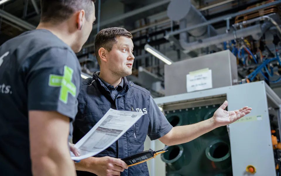 THE team discusses customer-specific concepts Two men in an industrial setting are conversing in front of a large machine unit, with one holding a document and the other making an explanatory hand gesture, surrounded by pipes and structural cabling.