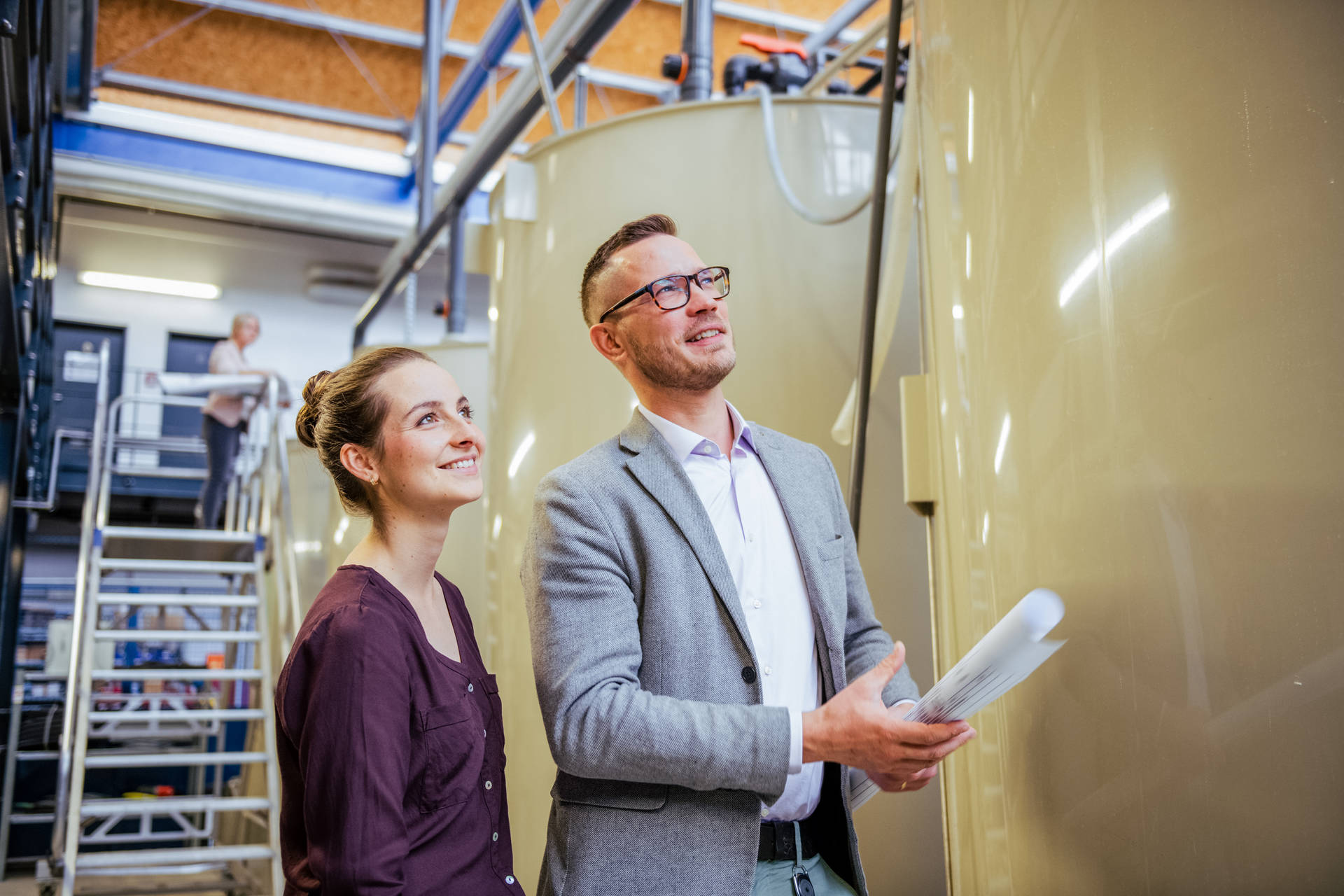 Two people in a modern, industrial facility stand smiling in front of large, beige tanks, holding papers in their hands, while in the background a person works on a ladder.