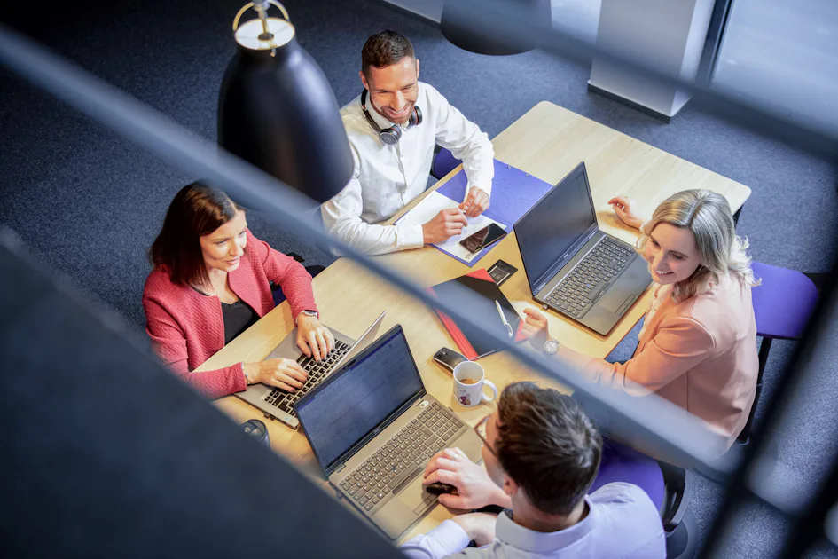 Press releases A group of four business professionals engages in a collaborative meeting around a wooden table, each working on laptops, with office furnishings and a window creating a modern open-plan workspace ambiance.