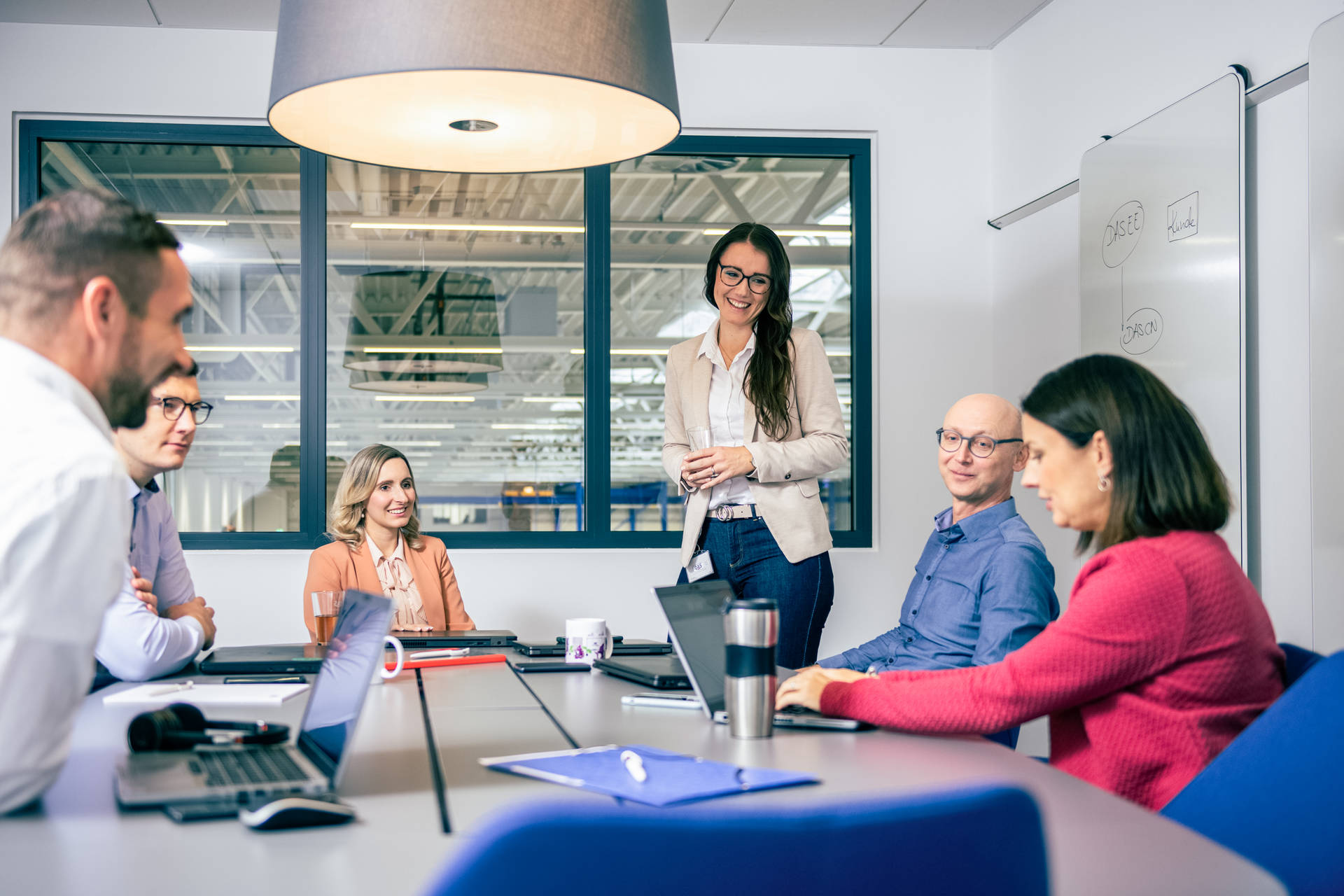 A group of professionals engaged in a meeting around a conference table in a modern office, with laptops and notebooks, surrounded by large windows and a hanging light fixture.