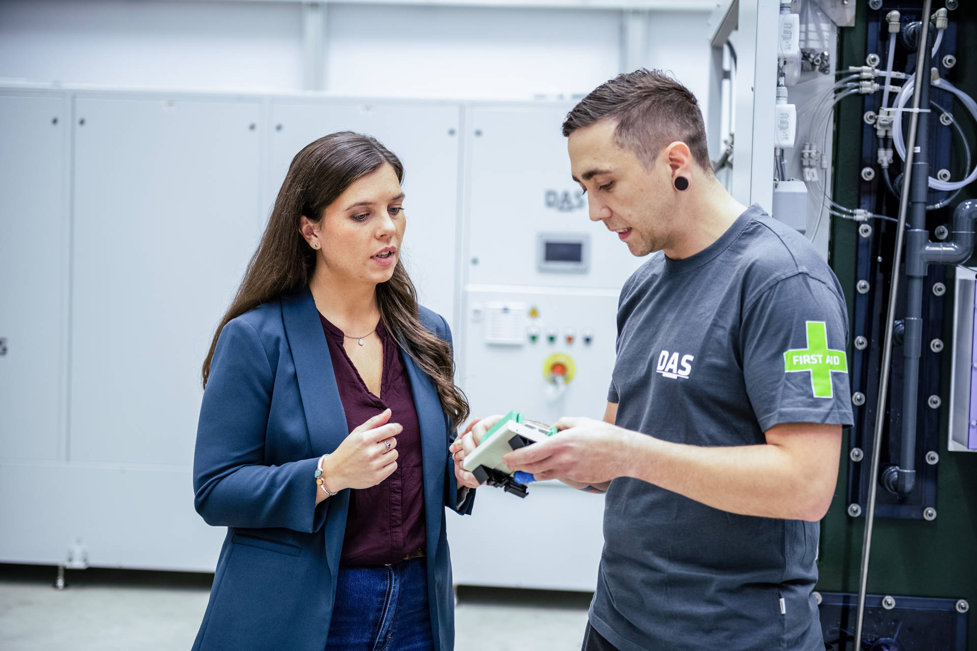 A woman and a man are standing in front of a large industrial plant, while the man is holding a technical device to talk about details.
