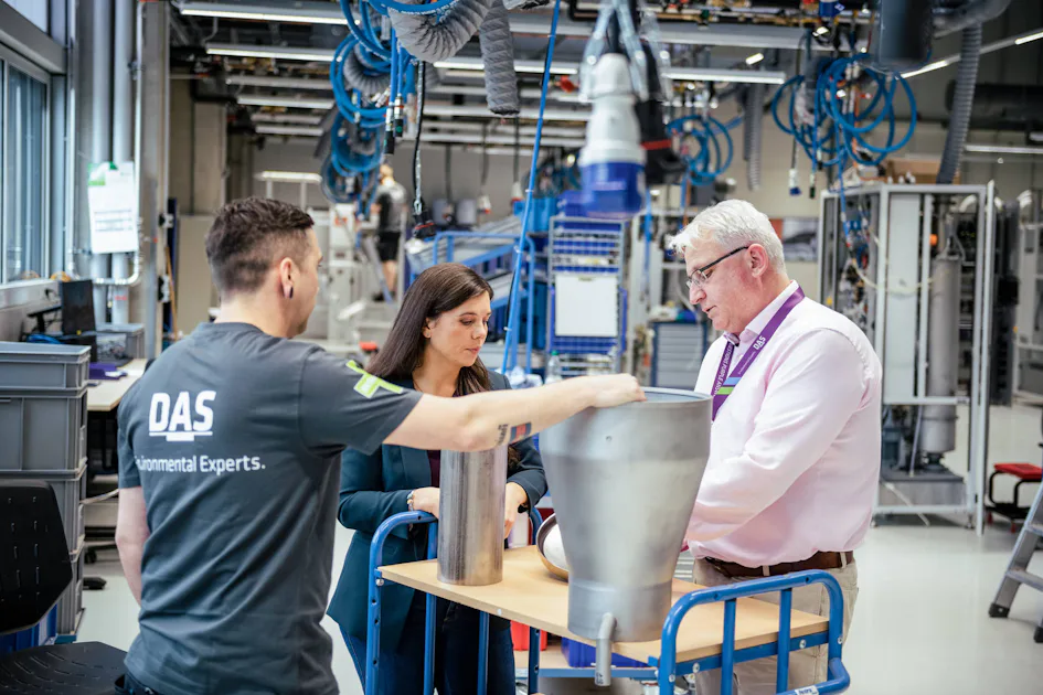 Our culture of personal development A group of three people are engaged in a discussion around industrial metal components on a table in a high-tech manufacturing facility filled with advanced machinery and blue cable systems.