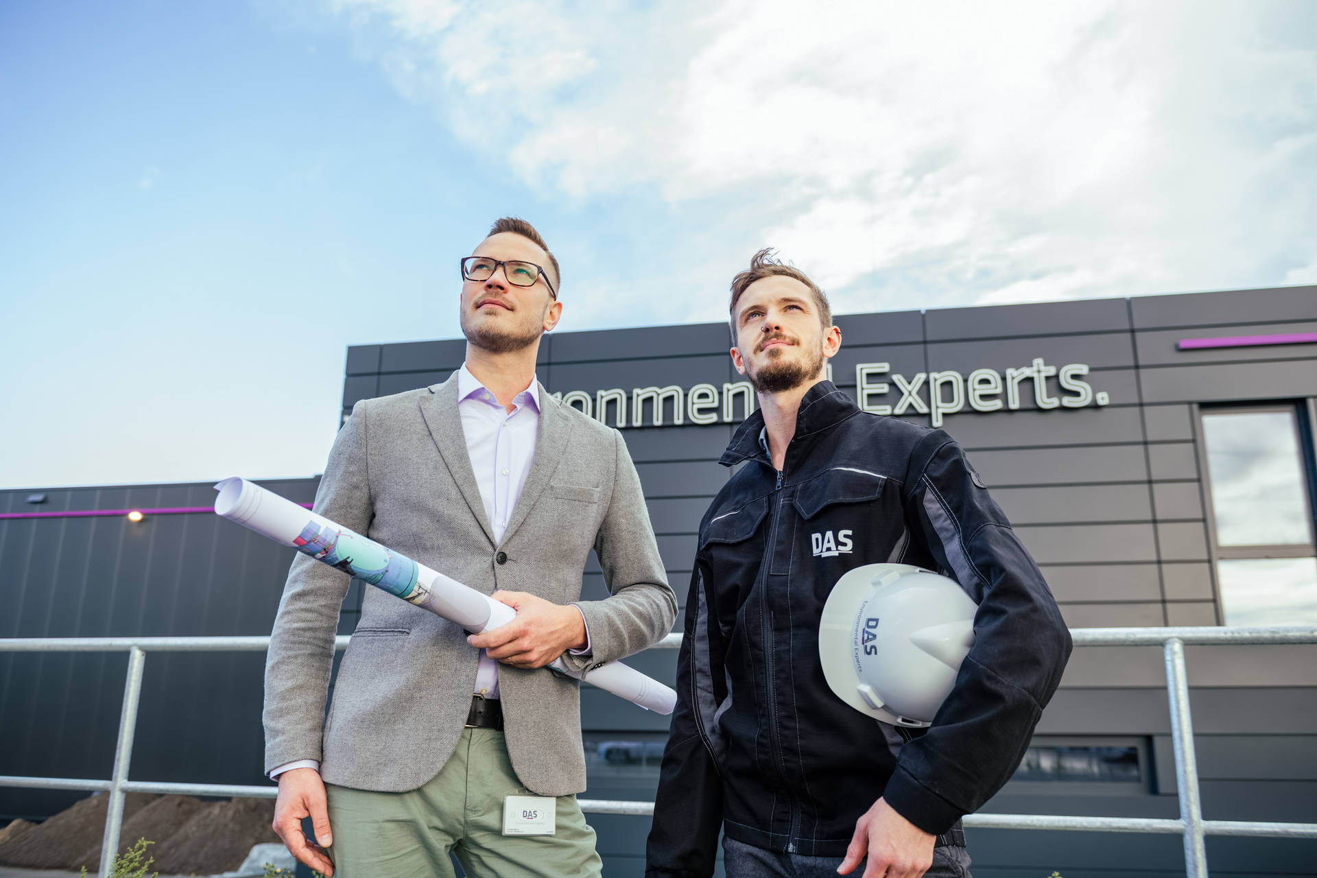 Two men are standing in front of a modern company building with the sign ‘Experts’. One is holding construction plans and the other a safety helmet with the inscription ‘DAS’.