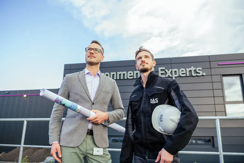 DAS Employee BUW 531 Two men are standing in front of a modern company building with the sign ‘Experts’. One is holding construction plans and the other a safety helmet with the inscription ‘DAS’.