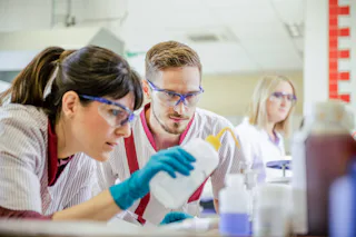 Laboratory staff analysing wastewater samples in the technical laboratory Three laboratory technicians wearing safety goggles and white coats closely examine a liquid in a bottle in a bright, modern laboratory setting.