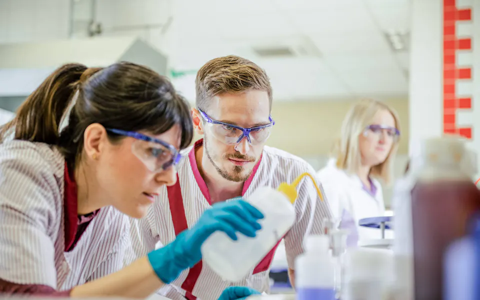 Laboratory staff analysing wastewater samples in the technical laboratory Three laboratory technicians wearing safety goggles and white coats closely examine a liquid in a bottle in a bright, modern laboratory setting.