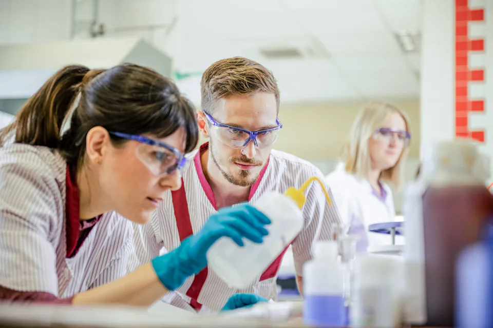 Three laboratory technicians wearing safety goggles and white coats closely examine a liquid in a bottle in a bright, modern laboratory setting.