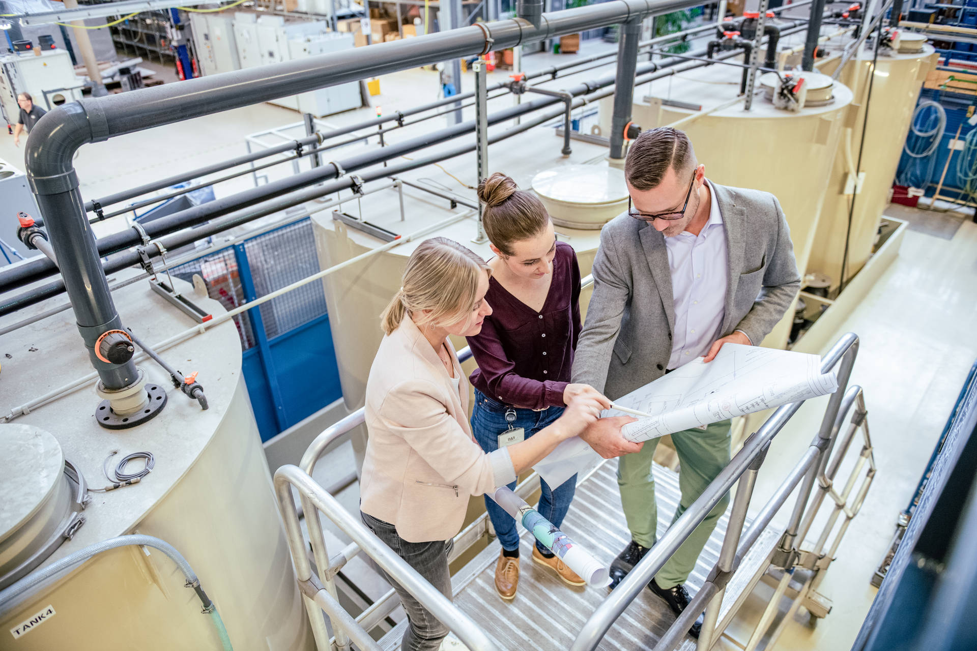 Three people stand on a metal staircase in a production hall and look at technical plans together, surrounded by industrial equipment and machinery.