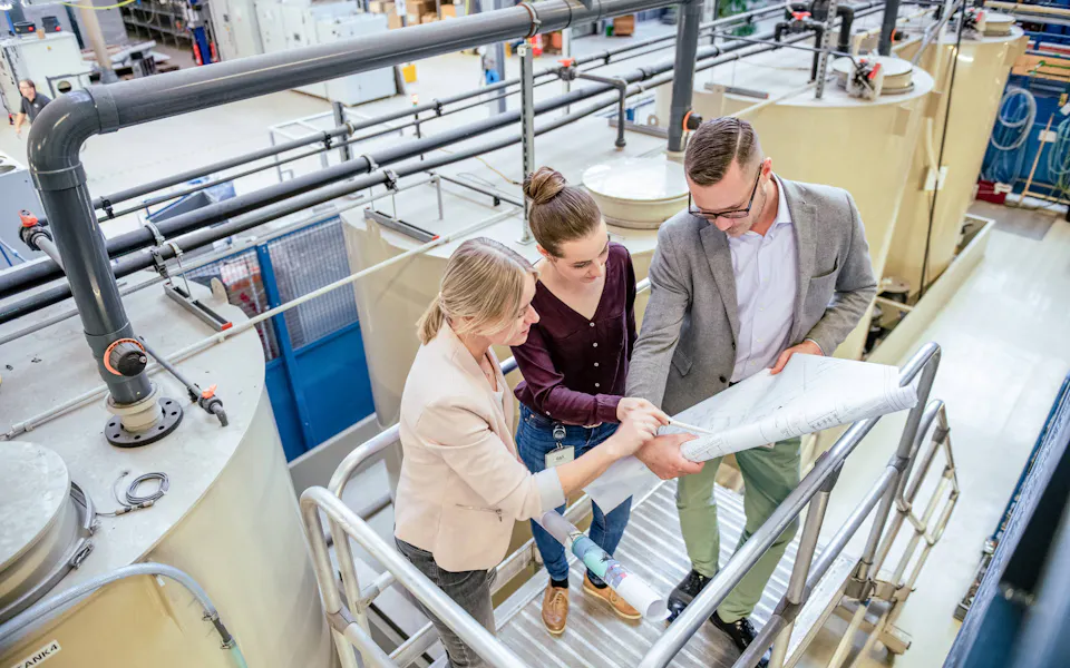 DAS 환경 전문가들이 폐수 처리 시설을 계획하고 있습니다. 세 사람이 생산 홀의 금속 계단에 서서 산업 장비와 기계류에 둘러싸인 채 기술 도면을 함께 살펴보고 있습니다.