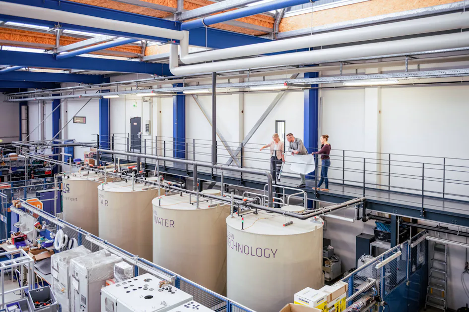 DAS EE Plant Construction Three people examine a large blueprint on an elevated walkway inside a modern industrial facility, with prominent tanks labeled “AIR,” “WATER,” and “TECHNOLOGY” positioned below.