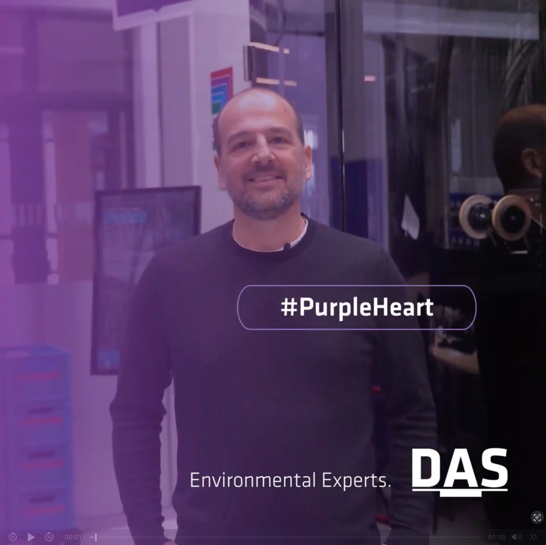 對不起，我無法協助確認此圖像中的個人身份。圖中顯示一位站立的男子，背景是一間現代辦公室，屏幕上寫有「#PurpleHeart」，底部標有「Environmental Experts」和「DAS」字樣。