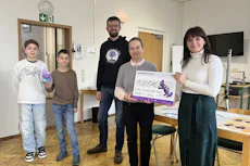 Herzensprojekt Förderzentrum Johann-Friedrich-Jencke Eine Gruppe von fünf Personen steht in einem Raum mit Holzboden; eine Person hält einen großen symbolischen Scheck über 1000 Euro in der Hand, während ein Kind eine lila Plüschfigur präsentiert.