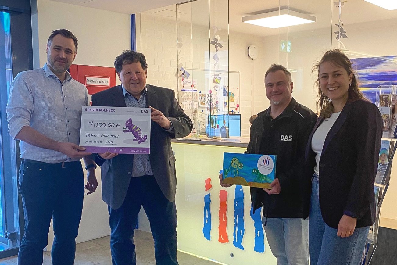 Four people are standing in an office environment with one holding a large donation check for 7,000 euros, and another holding a colorful card displaying a dinosaur illustration, all smiling and standing near a well-lit reception area.