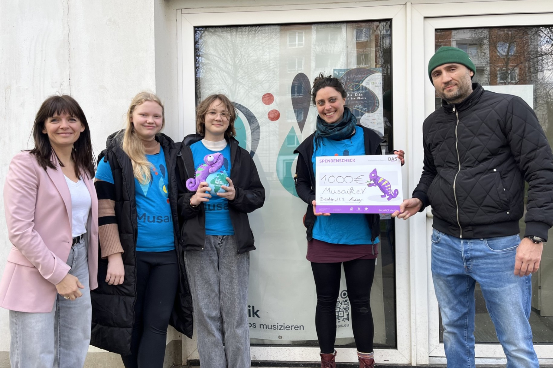 A group of five people stands in front of a building entrance, three of them holding a large donation check and a seahorse toy, all smiling and dressed casually.