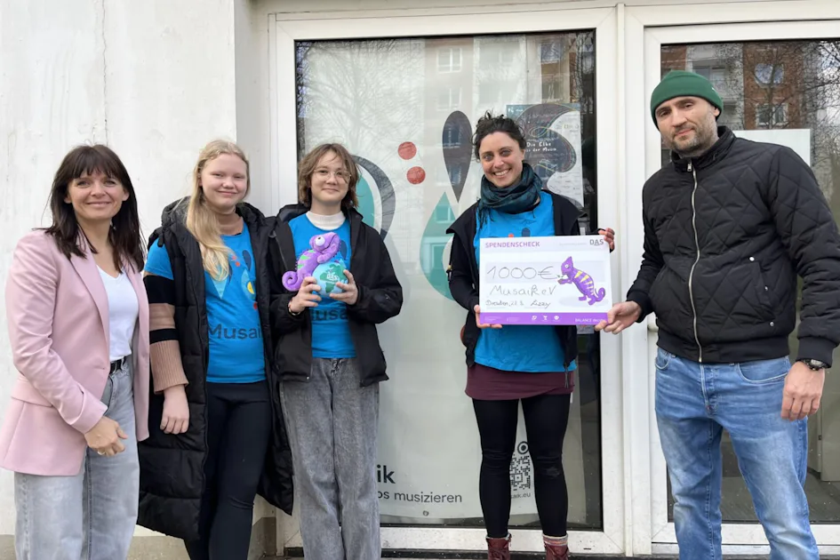 Herzensprojekt: Musaik Fünf Personen stehen vor einem Gebäude, zwei halten einen großen Scheck mit der Aufschrift "10,000€" und einem gezeichneten Seepferdchen, während die anderen eine Trophäe in Form eines Seepferdchens präsentieren.