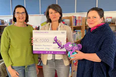 Herzensprojekt: Dresden Placetobe!. e.V. In einer Bibliothek überreichen drei Frauen einen Spendenscheck über 1000 Euro mit einem bunten Chamäleon-Design, während eine Frau ein lila Stoffchamäleon hält.