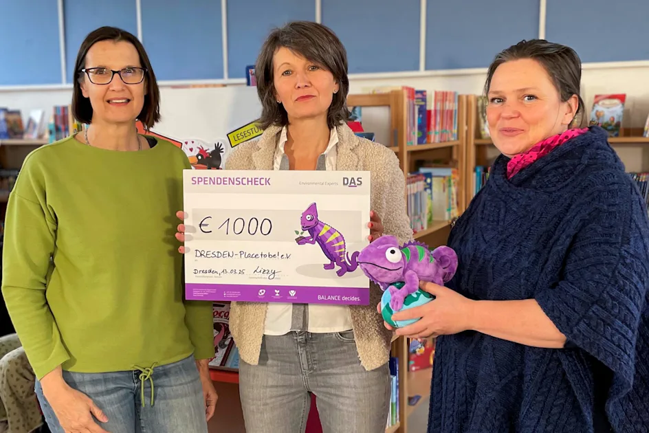 Herzensprojekt: Dresden Placetobe!. e.V. In einer Bibliothek überreichen drei Frauen einen Spendenscheck über 1000 Euro mit einem bunten Chamäleon-Design, während eine Frau ein lila Stoffchamäleon hält.