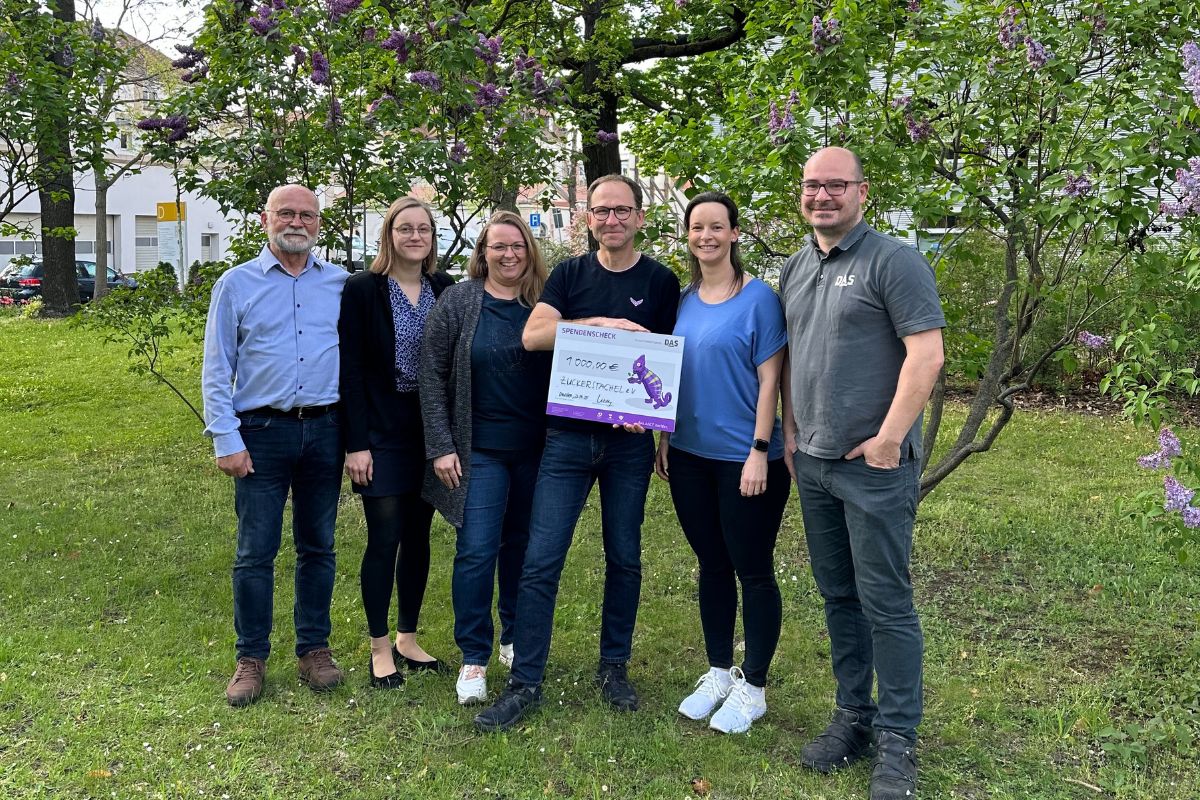 A group of six people stand together outdoors in a grassy area, holding a large sign with text, surrounded by blooming lilac bushes and trees in the background.