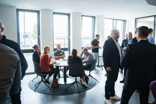 2. Industriewassertag In einem modernen, gut beleuchteten Büroraum stehen mehrere Personen im Gespräch, während eine Gruppe an einem Tisch sitzt und diskutiert, mit großen Fenstern im Hintergrund, die natürliches Licht hereinlassen.