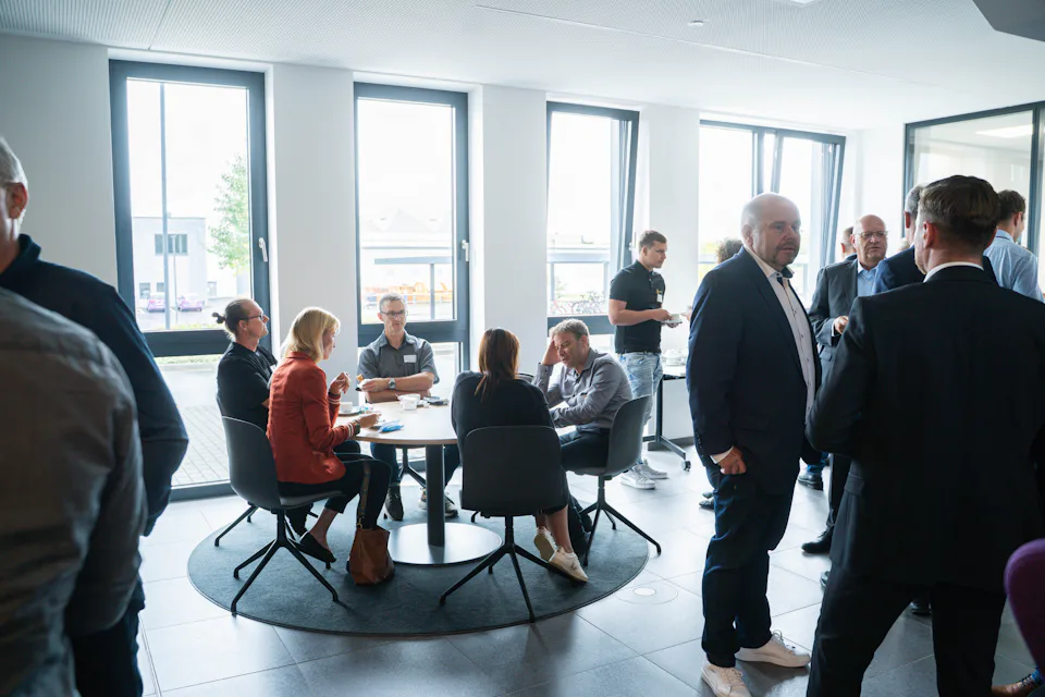A group of people are engaged in conversation and networking around a table in a bright, modern office with large windows and natural light.