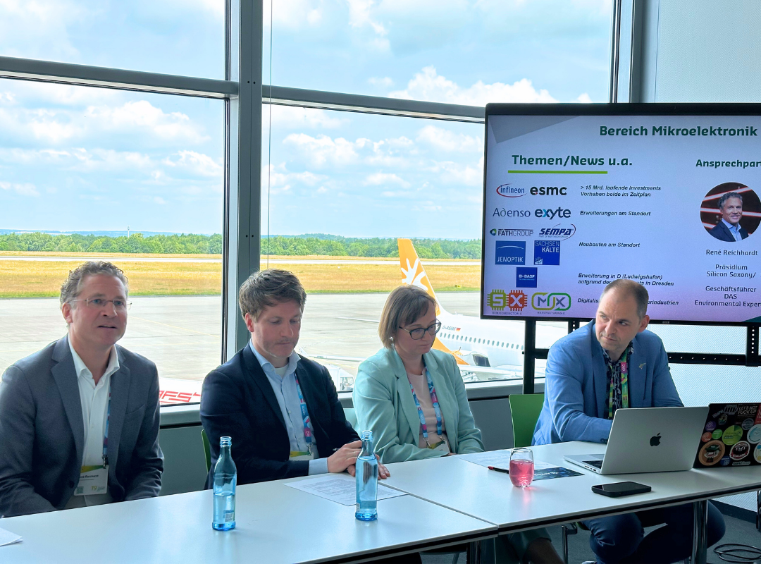 Eine Gruppe von vier Personen sitzt vor einem Tisch mit Laptop und Wasserflasche bei einer Präsentation über Mikroelektronik in einem Raum mit großen Fenstern und Blick auf das Rollfeld eines Flughafens.