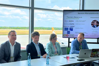19. Silicon Saxony Day Eine Gruppe von vier Personen sitzt vor einem Tisch mit Laptop und Wasserflasche bei einer Präsentation über Mikroelektronik in einem Raum mit großen Fenstern und Blick auf das Rollfeld eines Flughafens.