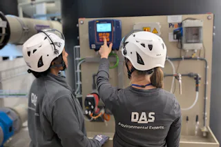 Vorletzter Meilenstein bei Okmetic erreicht Zwei Personen mit Schutzhelmen arbeiten an einem Kontrollpanel in einer industriellen Anlage und interagieren mit der Anzeigetafel, um die Umweltsysteme zu überwachen.