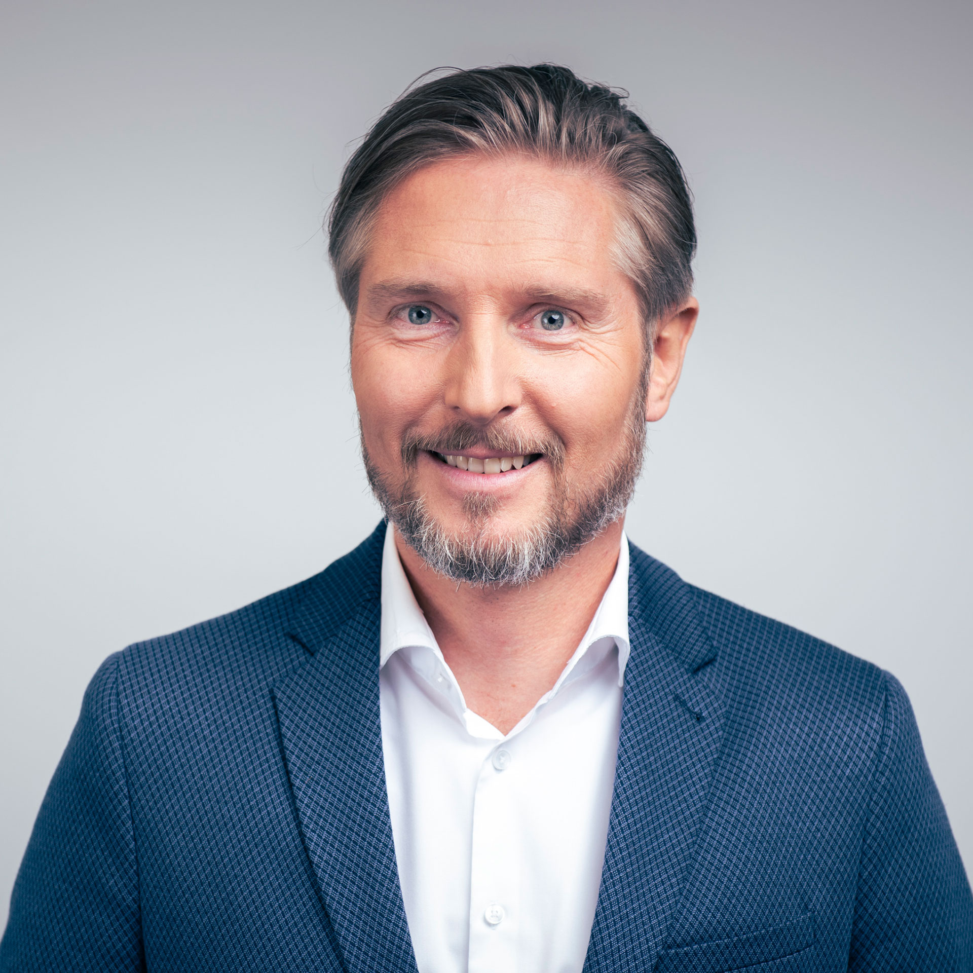 짧고 밝은 갈색 머리와 수염을 기른 ​​미소 띤 남자가 흰 드레스 위에 파란색 질감의 블레이저를 입고 있습니다.