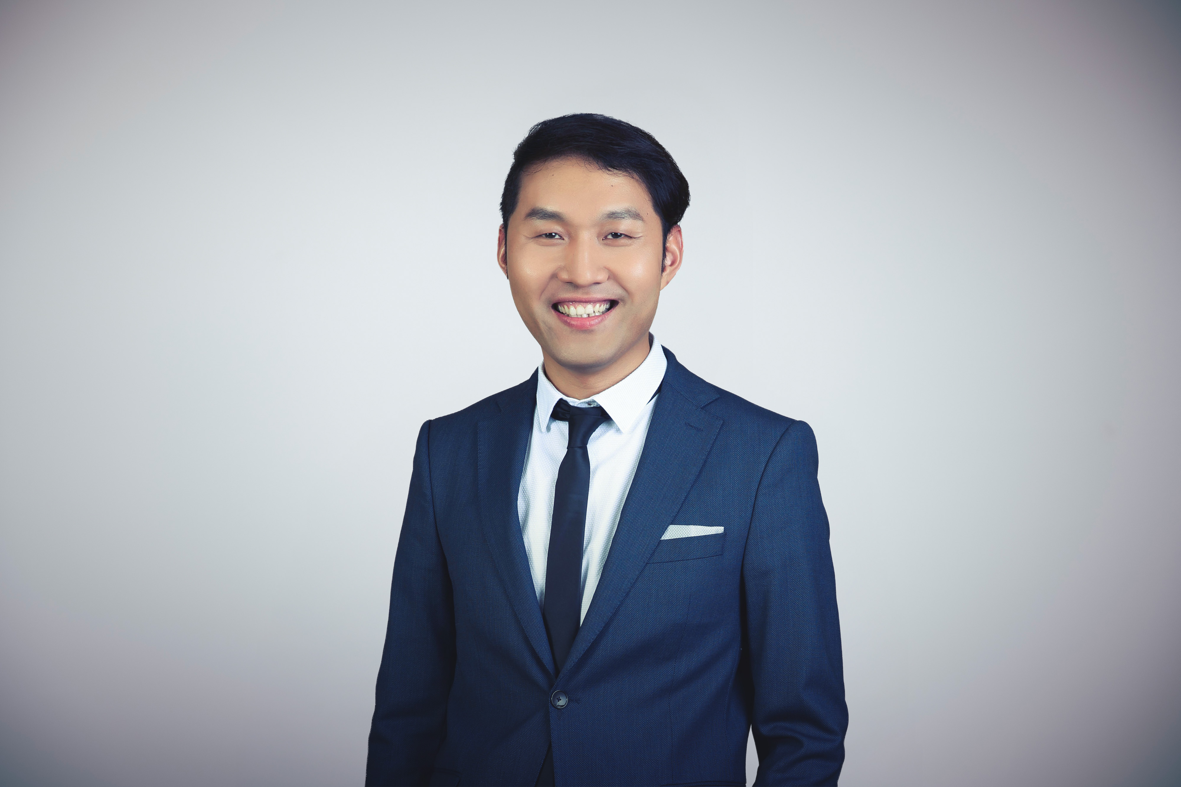 A person in a blue suit and tie smiles confidently against a plain white background, capturing a professional and approachable demeanor.