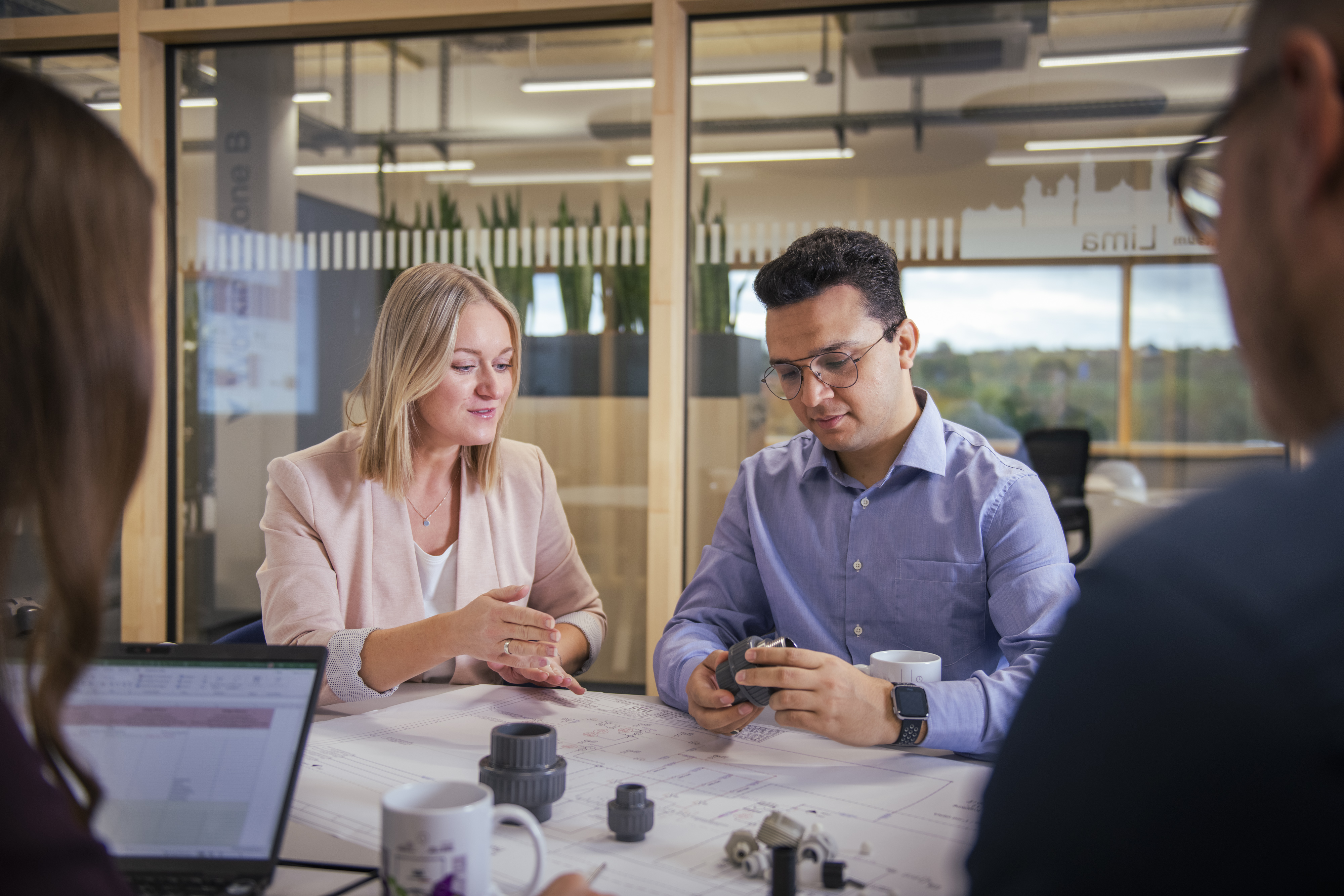 Four people are engaged in lively discussion at a table in a modern office, surrounded by models of machine components and laptops, in front of a glass wall with green plants in the background.