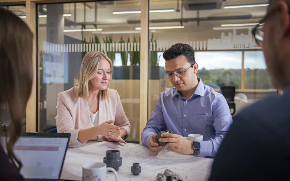 The EE Team plans new plant Four people are engaged in lively discussion at a table in a modern office, surrounded by models of machine components and laptops, in front of a glass wall with green plants in the background.