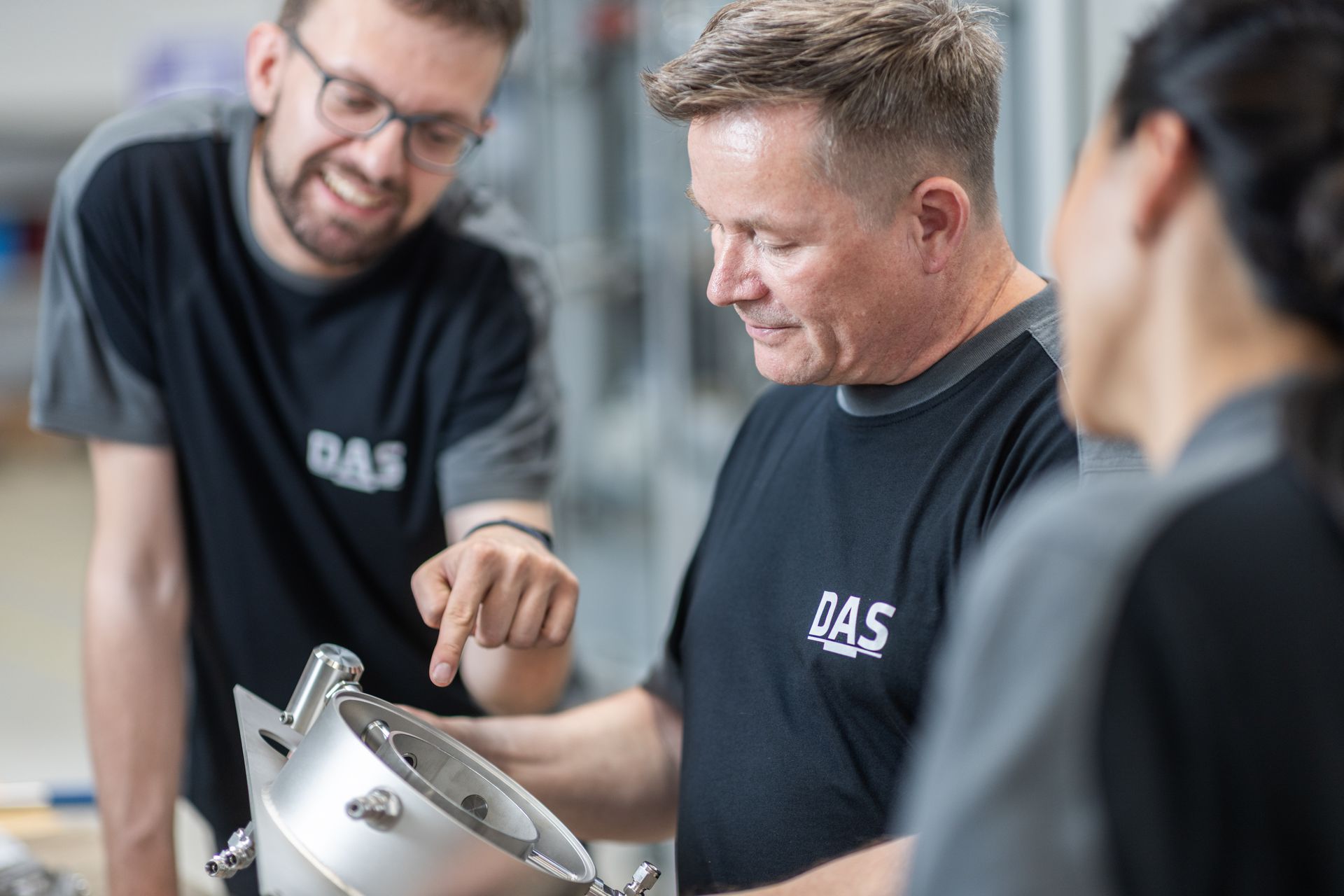 Three people, wearing matching DAS-logo black shirts, are gathered around an industrial mechanical component, with one individual pointing at its features while discussing it enthusiastically in a well-lit workshop.