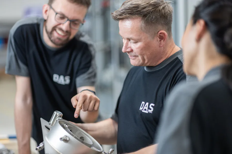 Certified Manufacturing Quality at DAS in Dresden/Germany Three people, wearing matching DAS-logo black shirts, are gathered around an industrial mechanical component, with one individual pointing at its features while discussing it enthusiastically in a well-lit workshop.