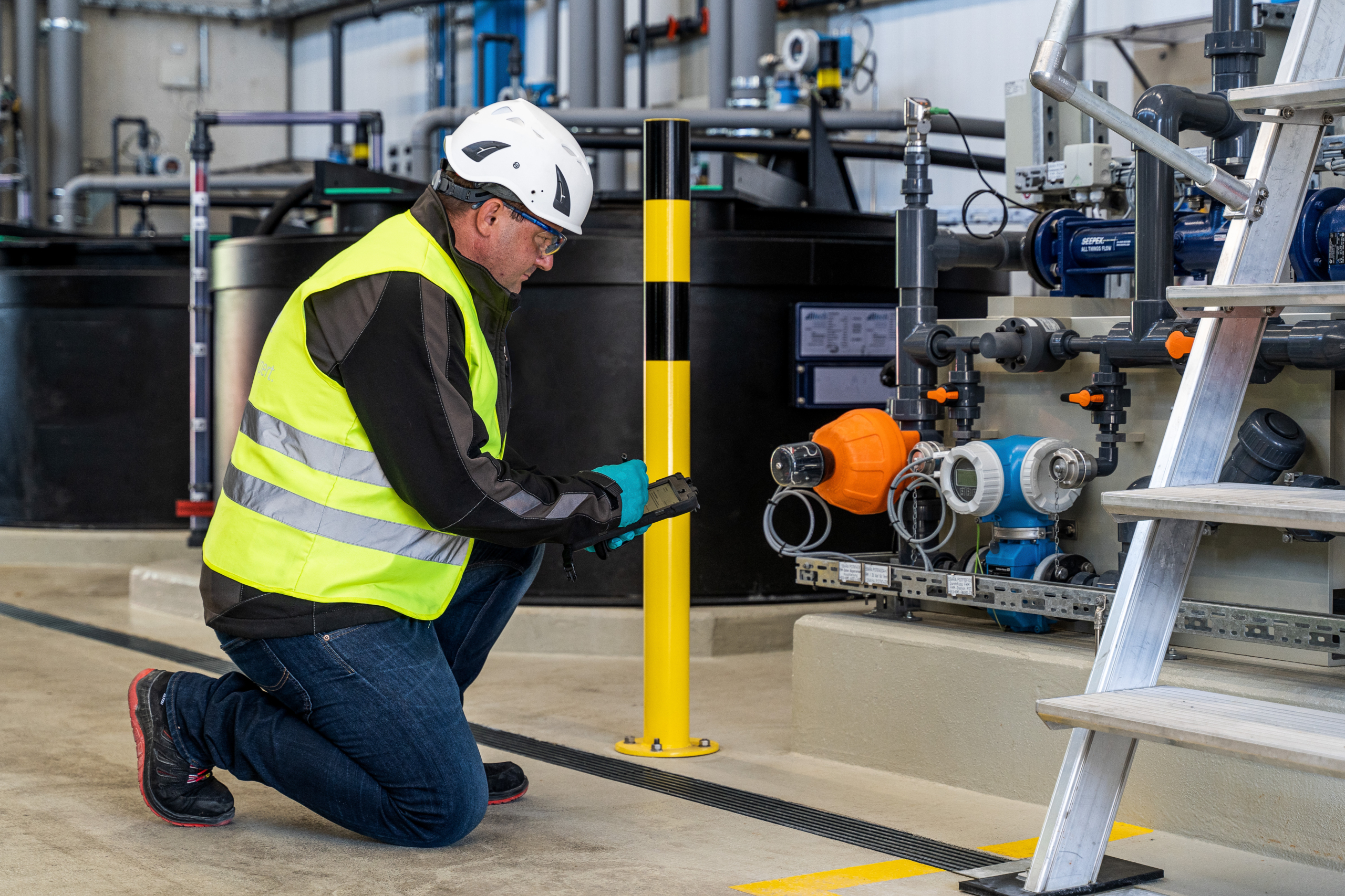 A worker in a safety vest and hard hat kneels beside an array of industrial machinery in an industrial plant, inspecting equipment and taking notes, emphasizing maintenance in a high-tech environment.