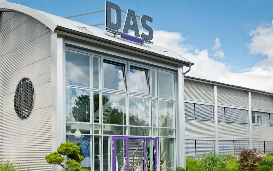 The Front of DAS Building A modern industrial building featuring large glass windows and a prominent gray and purple "DAS" sign on the rooftop, set against a partly cloudy sky.